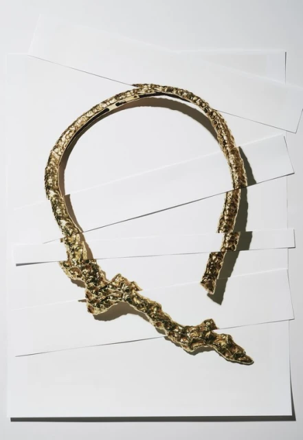 A gold collar necklace is artfully displayed against a background of layered white paper, highlighting its design and texture.