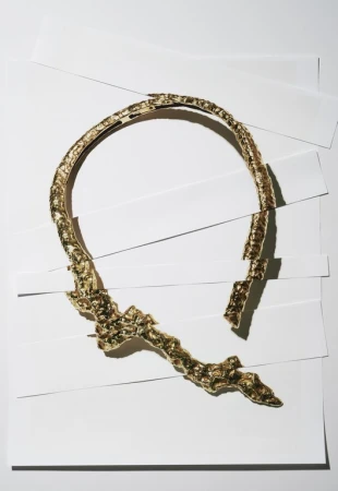 A gold collar necklace is artfully displayed against a background of layered white paper, highlighting its design and texture.