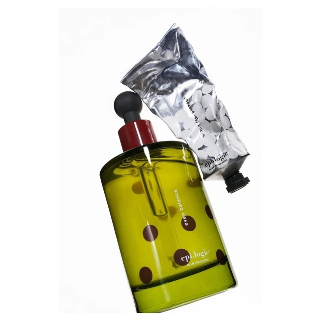 Cosmetic product still life featuring a green glass bottle with brown polka dots, a black dropper, and a silver tube on a white background.