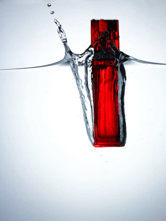 A red rectangular bottle causes a splash as it drops into clear water with a white background, creating dynamic water droplets and movement.