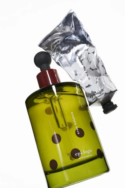 Still life of a bottle of 'Epilogue Even Balance' serum with brown polka dots and a tube of white cream on a white background.