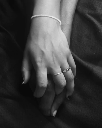 Monochrome close-up of two hands holding each other gently, wearing delicate rings and a thin bracelet against dark fabric.