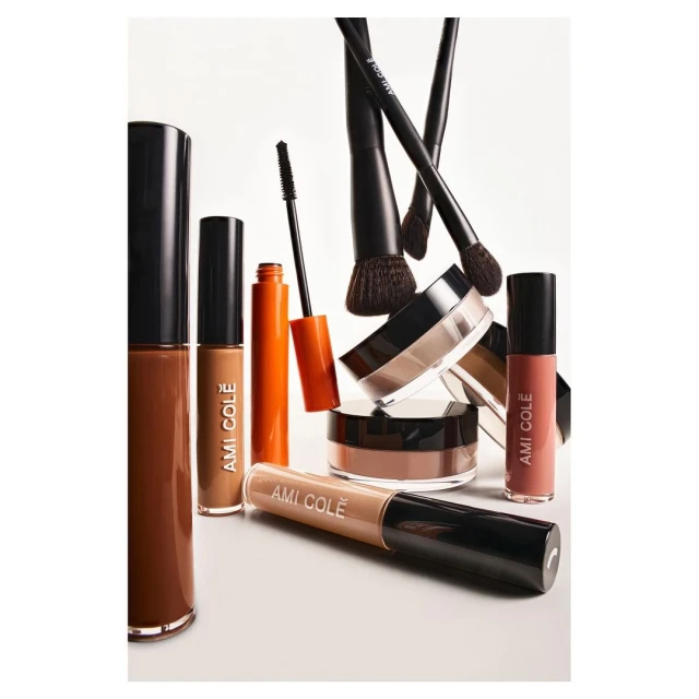 Cosmetic products from Ami Colé are displayed on a white surface. A collection of makeup brushes is visible in the background.
