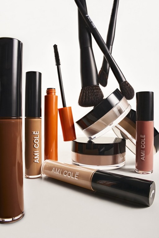 A collection of Ami Colé makeup products including foundation, concealer, powder, mascara, lip gloss, and makeup brushes against a white background.