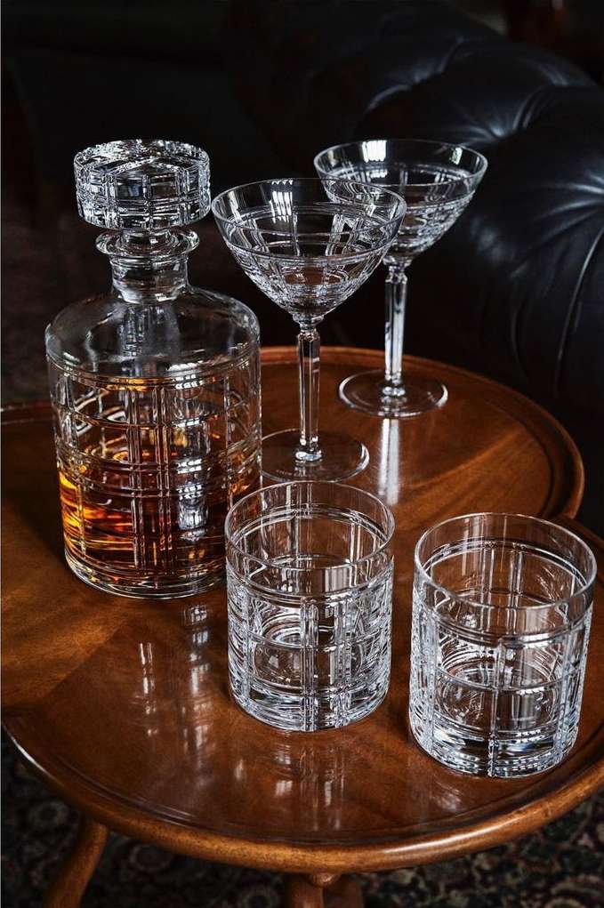 Crystal liquor decanter and glasses, including coupe and rocks styles, sit on a wooden side table against dark leather upholstery.