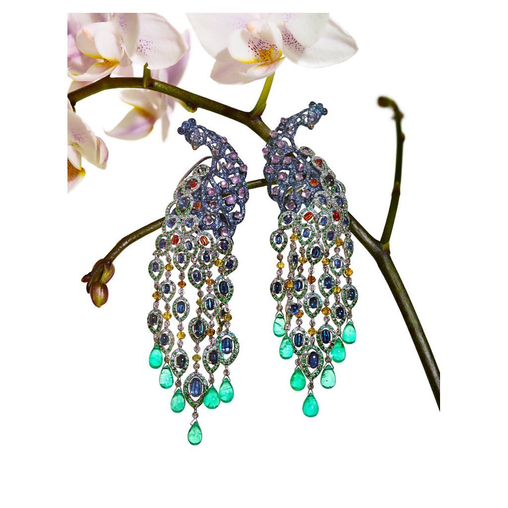 Close-up of ornate peacock earrings with colorful gemstones, set against a delicate white orchid and a stark white background. Jewelry.