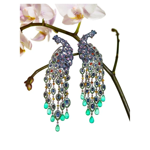 Close-up of ornate peacock earrings with colorful gemstones, set against a delicate white orchid and a stark white background. Jewelry.
