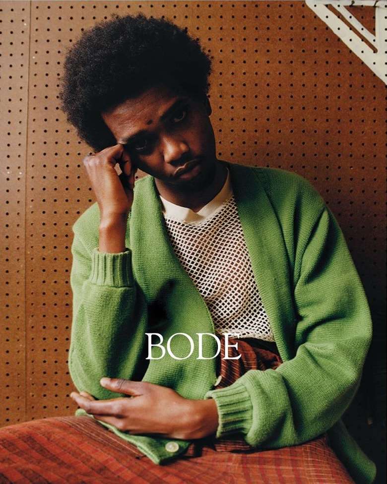 A portrait of a black male model in a green cardigan and reddish brown pants, posed against a speckled brown background, indoors.