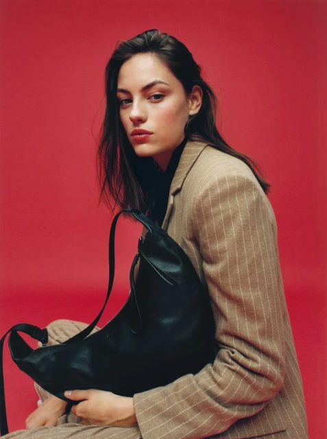 Adult female in a pinstripe suit holds a black leather bag against a red studio backdrop with a neutral expression and medium shot.