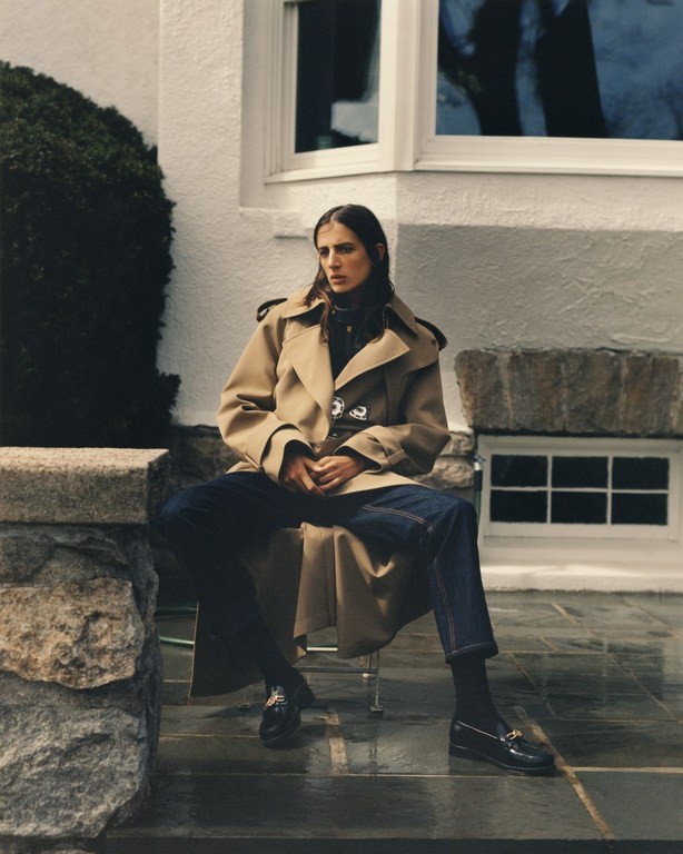 A stylish woman in a trench coat and jeans sits outdoors, complemented by elegant loafers and classic architecture in the background.