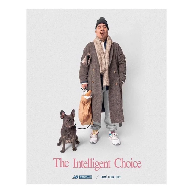 Man in coat and hat walking a French bulldog while carrying a paper bag with baguettes, studio shot against a plain white backdrop.