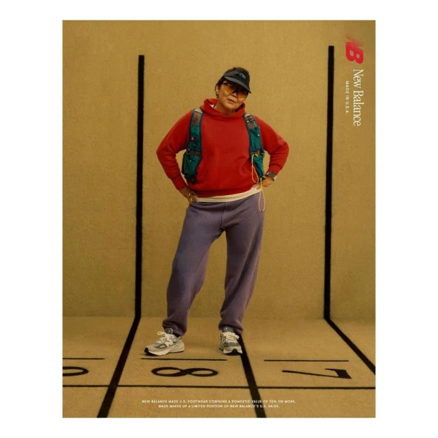 Woman in sportswear poses against a tan backdrop with track lines, showcasing an athletic red hoodie, lavender sweats, and New Balance sneakers.