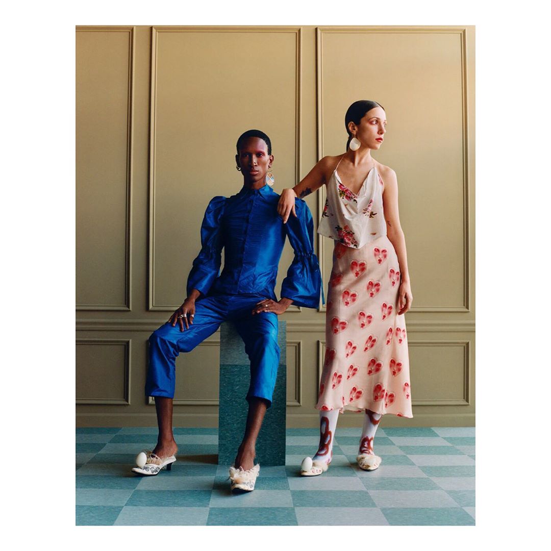Two people pose in a fashion editorial shot in a photo studio, wearing floral and blue outfits and unique footwear.