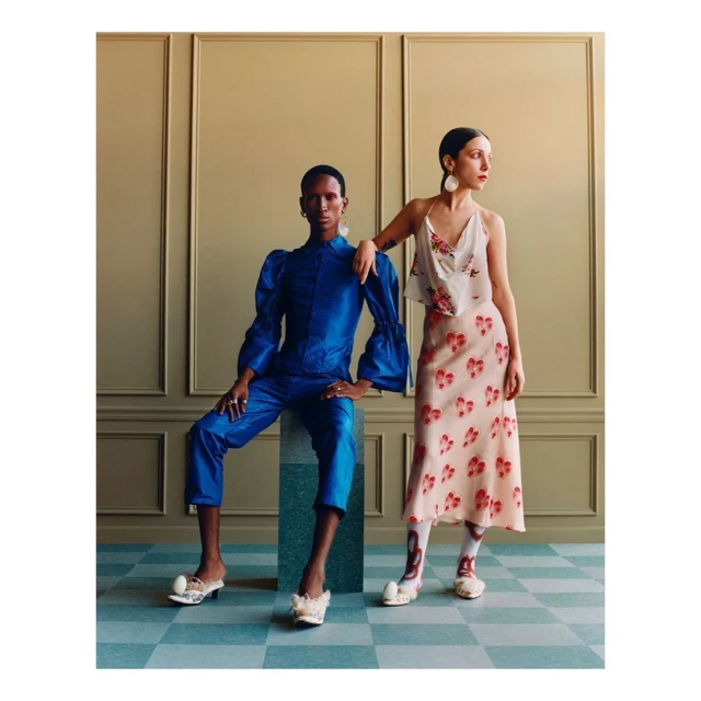 Two people pose in a fashion editorial shot in a photo studio, wearing floral and blue outfits and unique footwear.