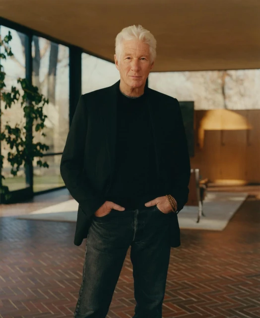 Portrait of an adult male with white hair, wearing a black blazer, black shirt, and jeans, posing indoors with natural window lighting.