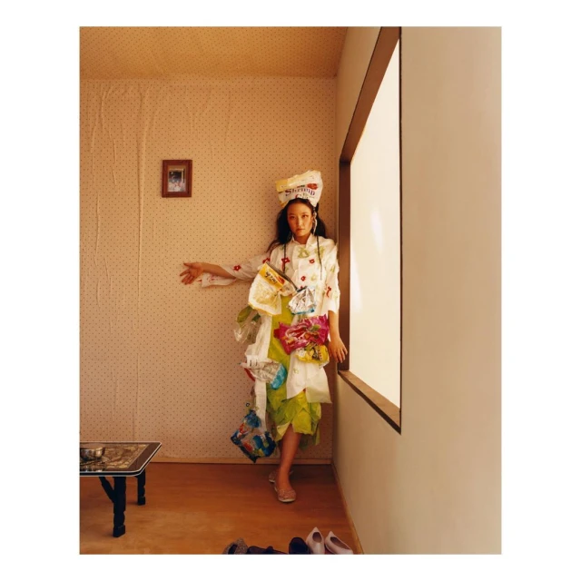 Conceptual fashion shot of an Asian woman in a paper hat and paper dress, standing in a room with natural light. Multiple handbags.