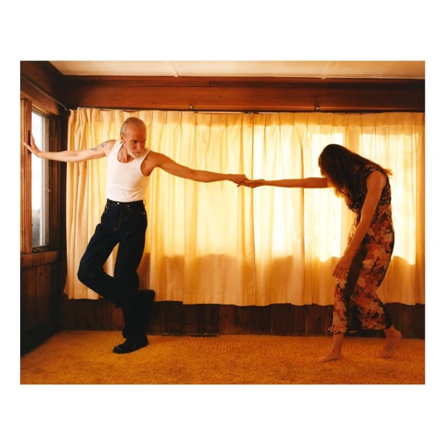 Couple dancing in a room, with a man in a white tank top and jeans and a woman in a floral dress holding hands.