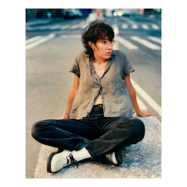 A young adult woman sits cross-legged on a concrete barrier with a street scene in the background, wearing casual clothing and white shoes.