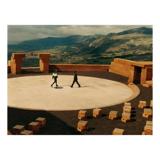 Two people walk across a circular stone stage surrounded by stone walls and block formations, with a mountain range in the background.