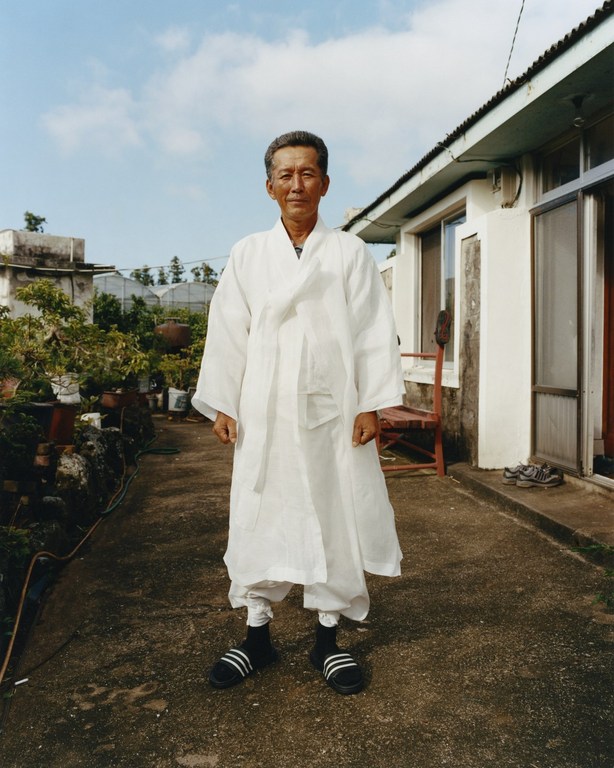Elderly Asian man in traditional white robe and modern sandals standing outdoors in front of house and garden on a sunny day.