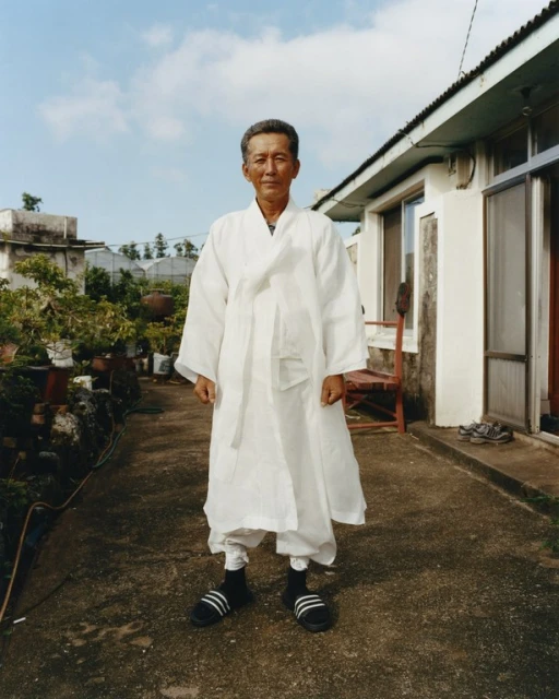 Elderly Asian man in traditional white robe and modern sandals standing outdoors in front of house and garden on a sunny day.