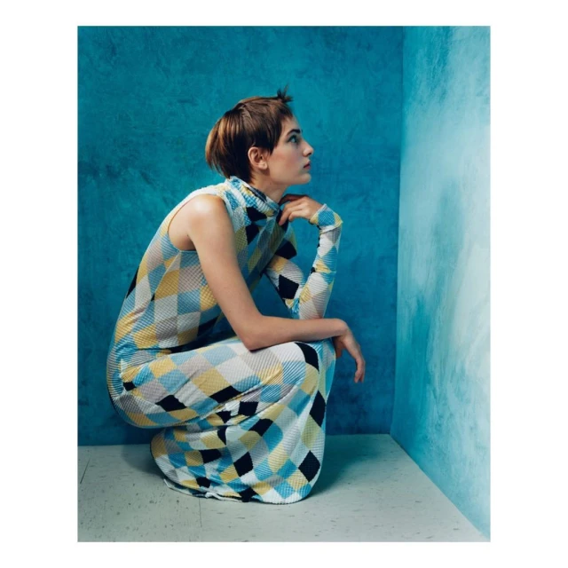A woman in a patterned dress is crouched in a corner against a mottled blue wall, looking up and to the side in a fashion pose.