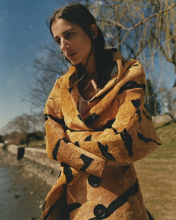A woman stands outside wearing a mustard yellow textured coat with abstract black stripes, on a sunny day with a river in the background.