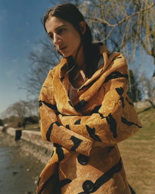 A woman stands outside wearing a mustard yellow textured coat with abstract black stripes, on a sunny day with a river in the background.