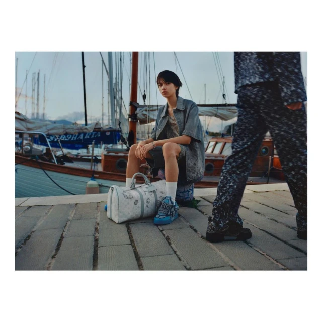 Young person in casual wear sits on a stone bollard by the water, a branded duffel at feet, boat masts rising in the soft-lit background.