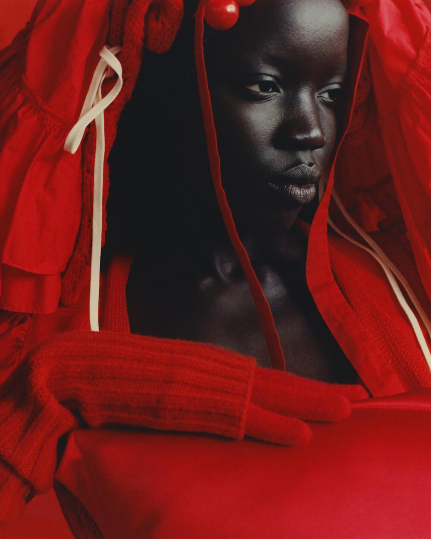 Portrait of a woman with dark skin wearing a red outfit and head covering, looking off to the side with a serious expression.
