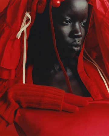 Portrait of a woman with dark skin wearing a red outfit and head covering, looking off to the side with a serious expression.