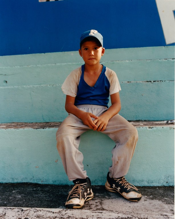 Young baseball player sits on concrete steps in a worn baseball uniform with sneakers, looking at the camera in this medium shot.
