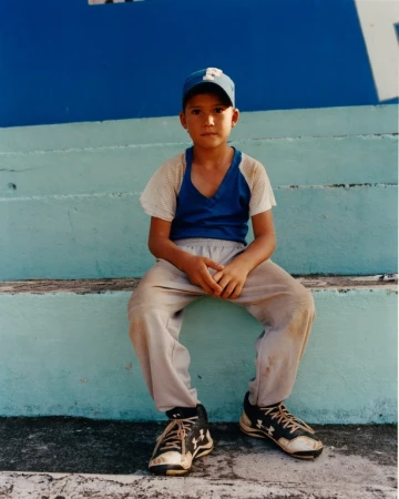 Young baseball player sits on concrete steps in a worn baseball uniform with sneakers, looking at the camera in this medium shot.