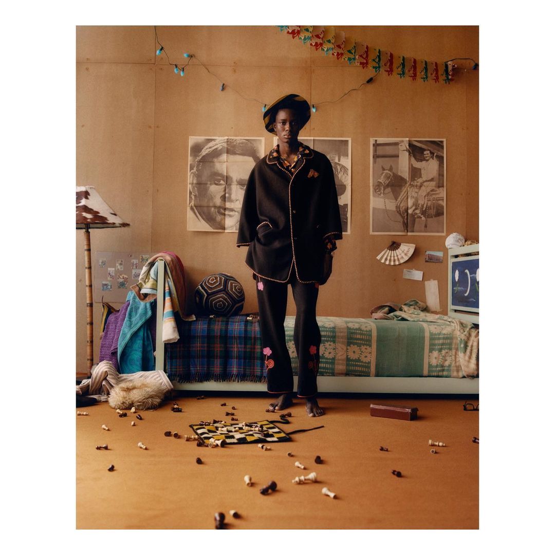 An African American woman in black pajamas and hat standing in a bedroom with a bed, chessboard, and posters on the wall.