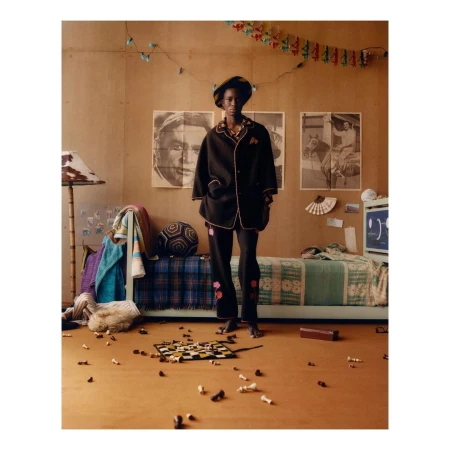 An African American woman in black pajamas and hat standing in a bedroom with a bed, chessboard, and posters on the wall.