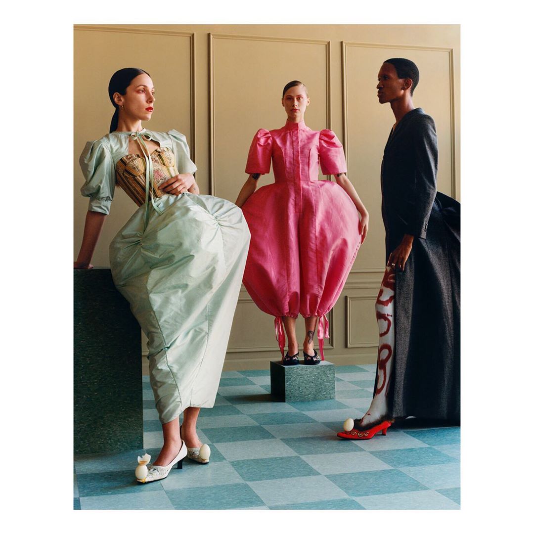 Three women in avant-garde dresses pose in a studio with a blue checkered floor, showcasing unique fashion and design elements.