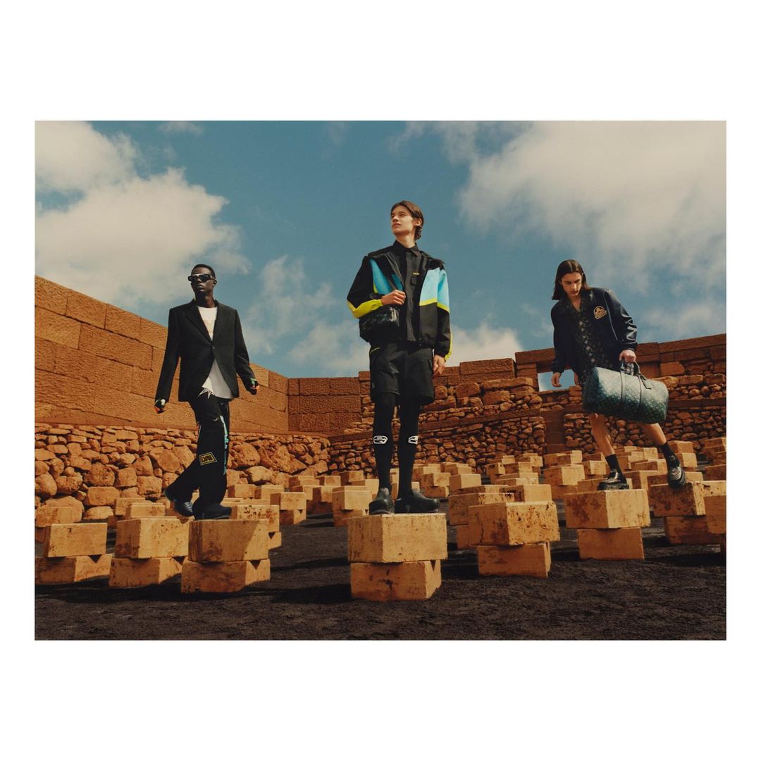 Three men in fashionable outfits stand on stone blocks in a barren landscape under a bright sky, showcasing a blend of modern style.