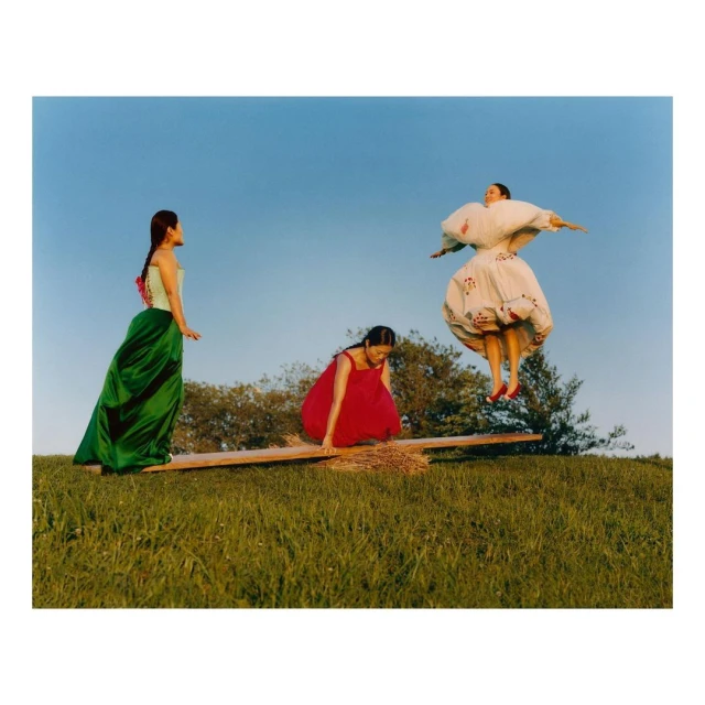 Three women in dresses on a grassy hill with a seesaw against a blue sky. One woman is jumping while the other two are on the ends.