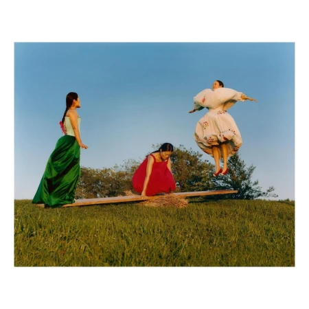 Three women in dresses on a grassy hill with a seesaw against a blue sky. One woman is jumping while the other two are on the ends.
