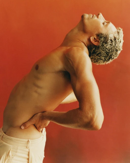 Shirtless man with white hair and tanned skin poses with hands on lower back against a warm red background in a studio environment.