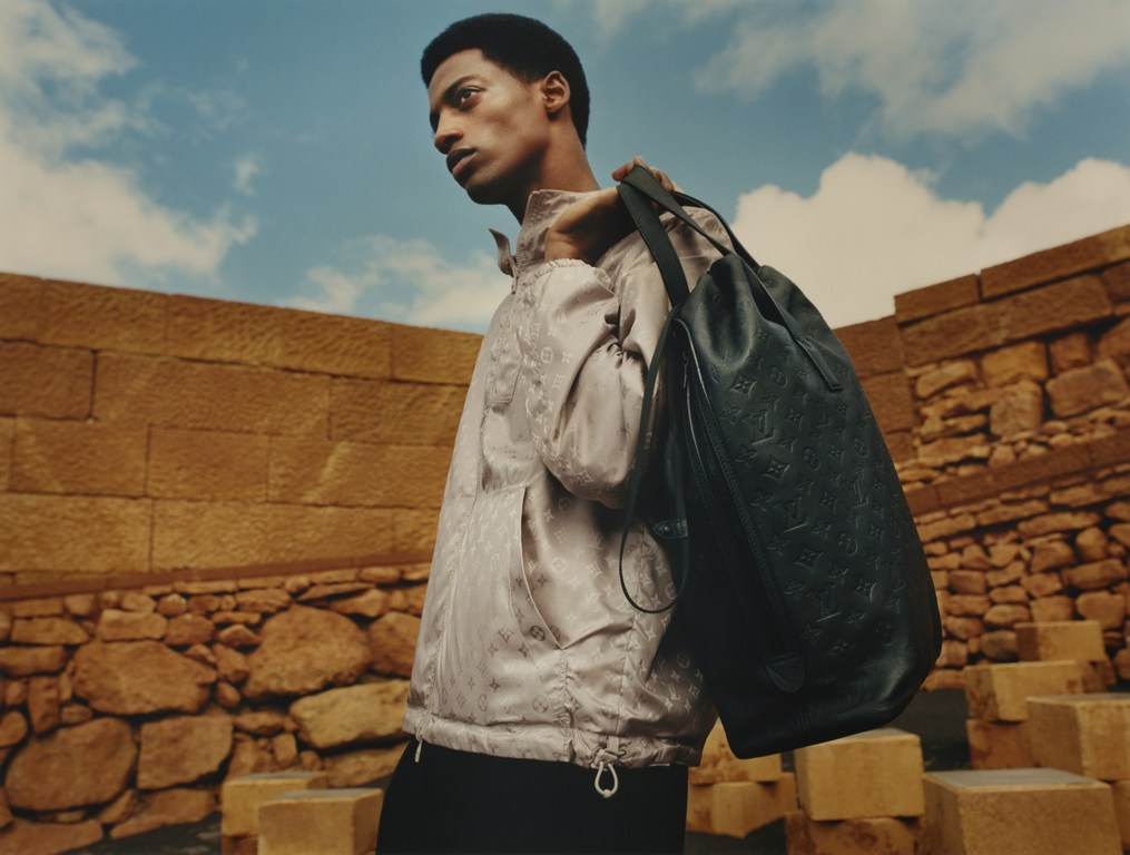 A man carries a black Louis Vuitton tote bag in front of a background of stone structures and a partly cloudy blue sky.
