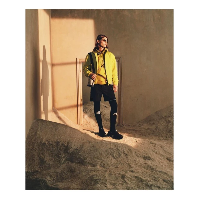 Athletic man stands on sandy dune in fashionable activewear: yellow hooded jacket, black shorts, knee-high logo compression socks, and sunglasses.