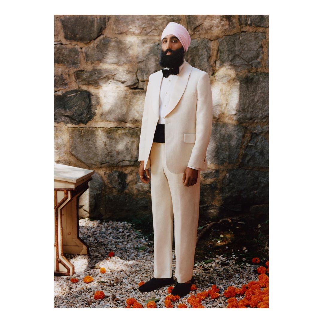 Man in white tuxedo and pink turban stands outdoors against stone wall with orange flowers on the ground in a medium, eye-level shot.