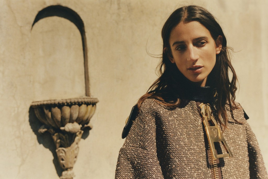 A young woman with long dark hair wearing a tweed jacket with an oversized gold zipper, posing in front of an architectural wall.
