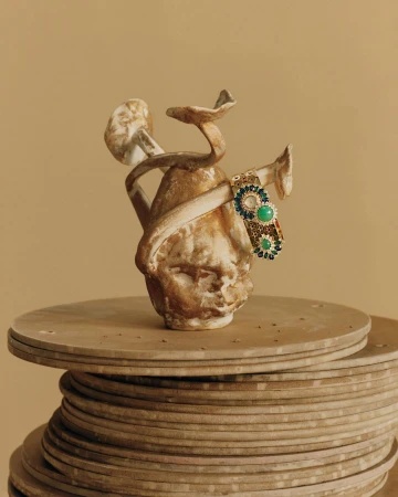 Still life shot of a ceramic head sculpture with mushroom-like growths adorned with a jeweled bracelet sitting on a tiered base.