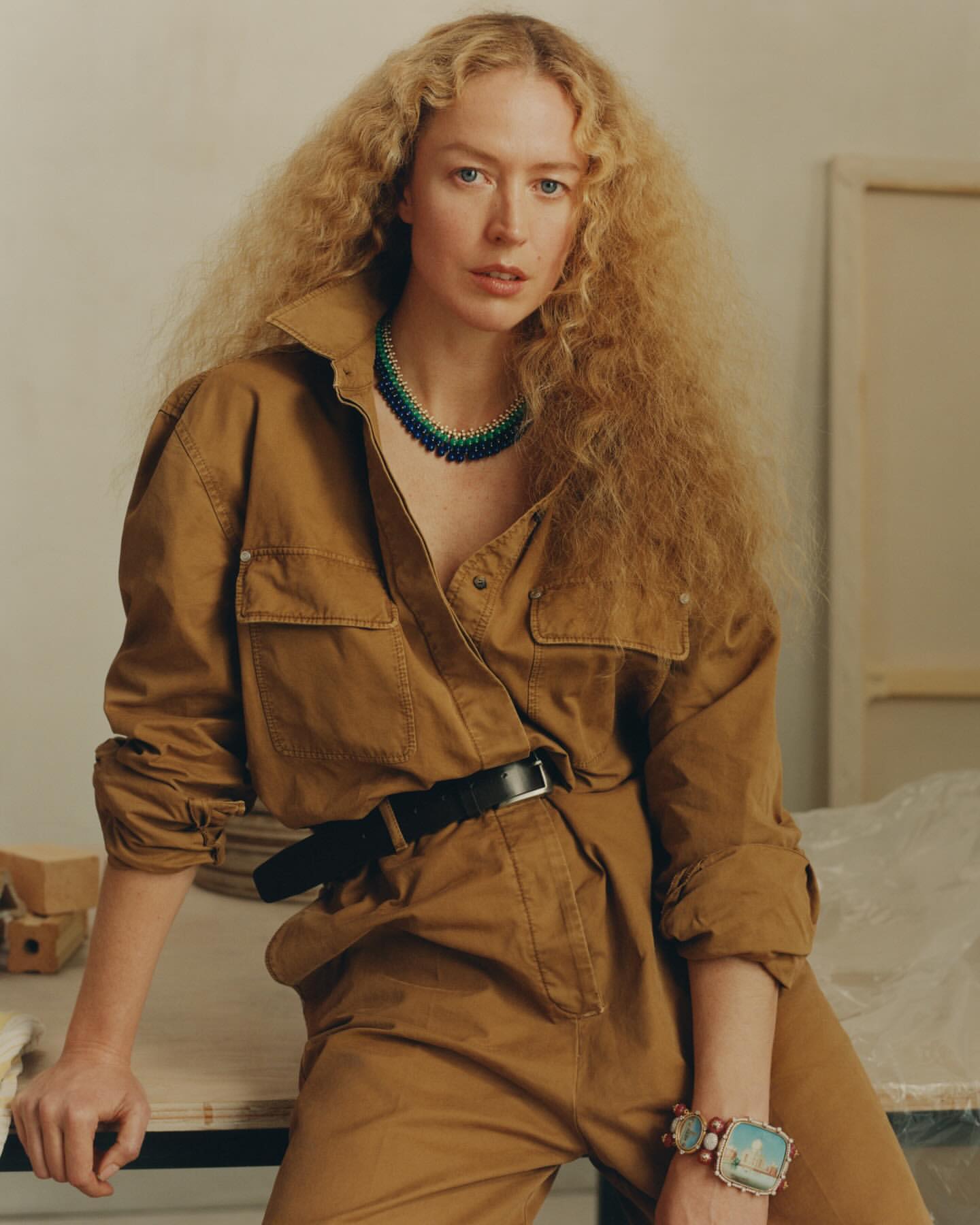 Adult woman with curly blonde hair poses in a tan jumpsuit with a black belt, wearing a green and blue necklace, in a studio setting.