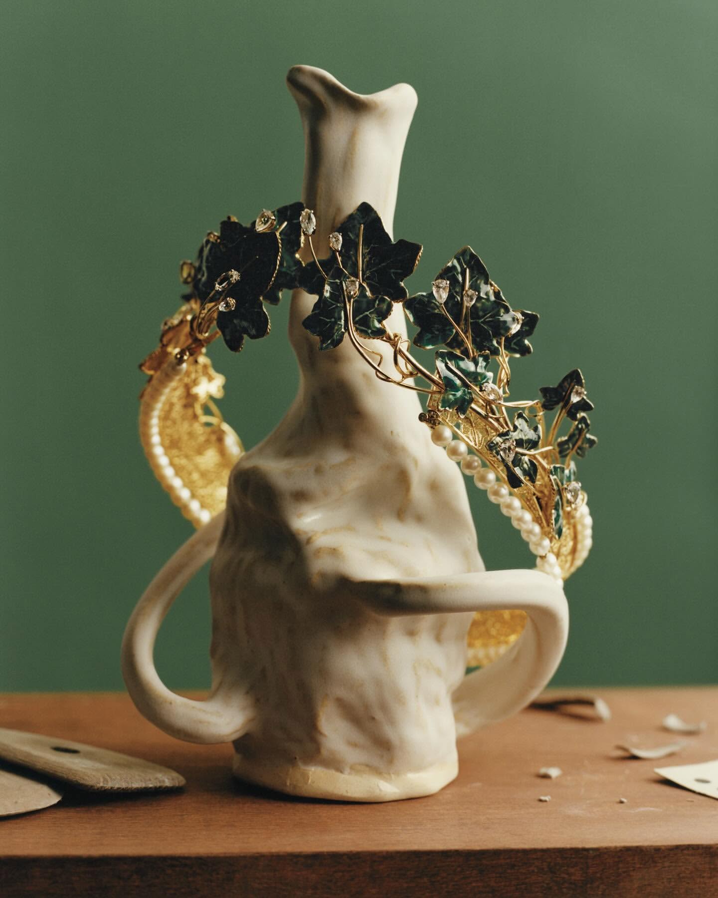 Still life of an off-white textured ceramic vase adorned with ornate ivy and pearl jewelry, set against a muted green background.