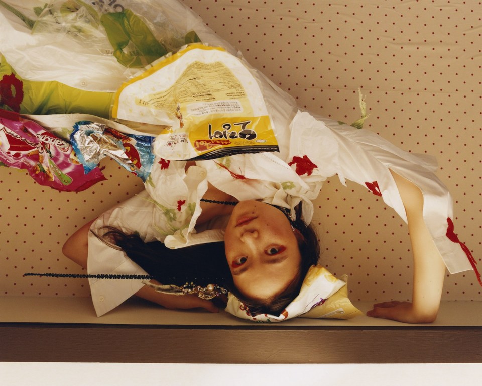 Asian woman lies beneath food wrappers and plastic bags, posing against a polka-dot background, creating an unconventional portrait.