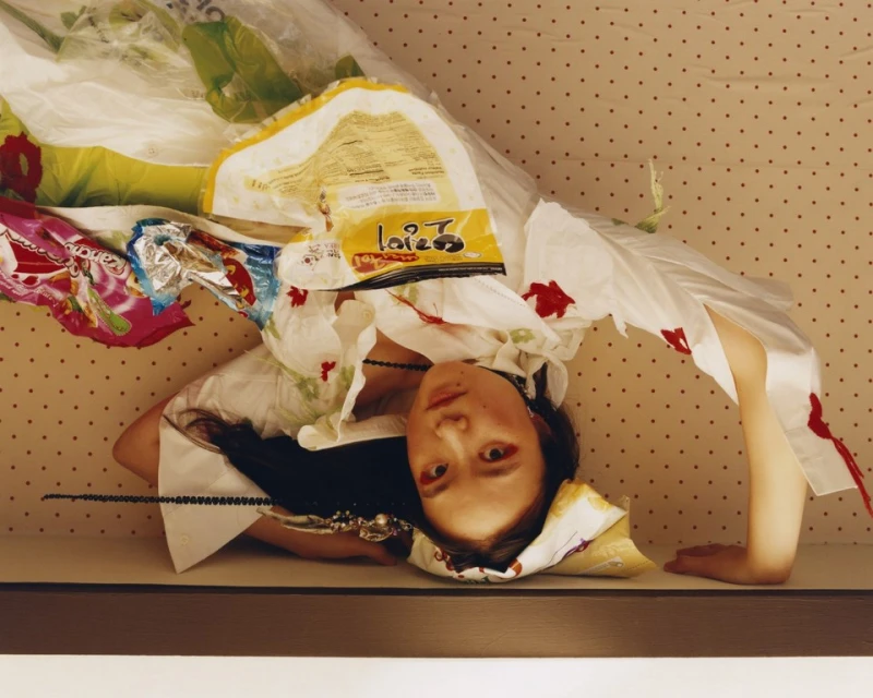 Asian woman lies beneath food wrappers and plastic bags, posing against a polka-dot background, creating an unconventional portrait.