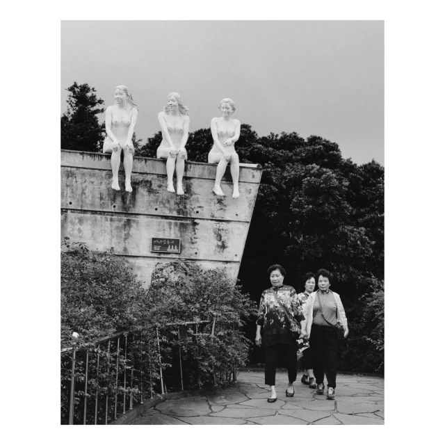 Black and white image of three nude statues perched on a concrete structure with women walking on a stone path below in a park.